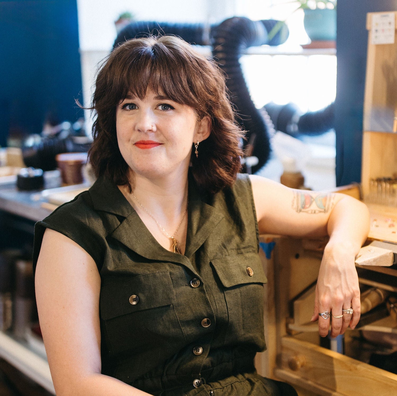 Jewelry artist Ali sitting in her workshop, wearing an olive green jumpsuit and showcasing a colorful tattoo on her upper arm. She has a confident expression, with jewelry tools and equipment visible in the background.