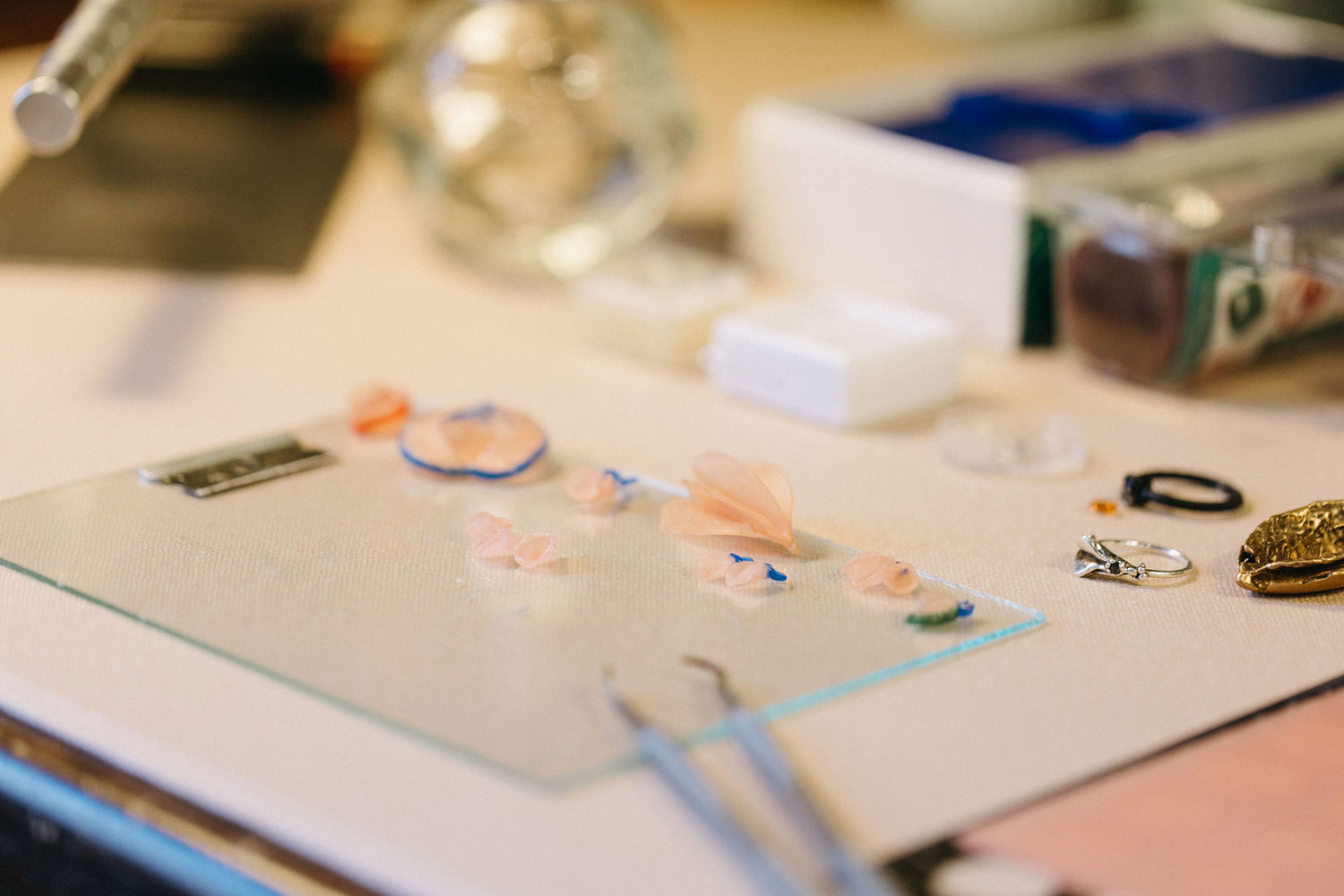 Close-up of a jewelry workspace with small pink and blue wax molds, tools, and rings arranged on a glass surface. The setup captures the intricate details of jewelry-making.