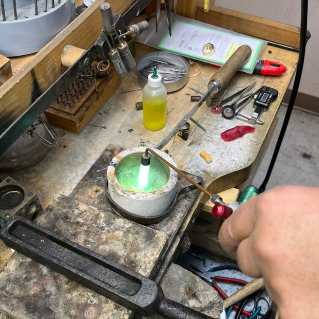 Jewelry workshop setup showing a melting crucible with green flame and a torch, surrounded by tools and materials. A hand holds a tool over the crucible in the metalworking process.